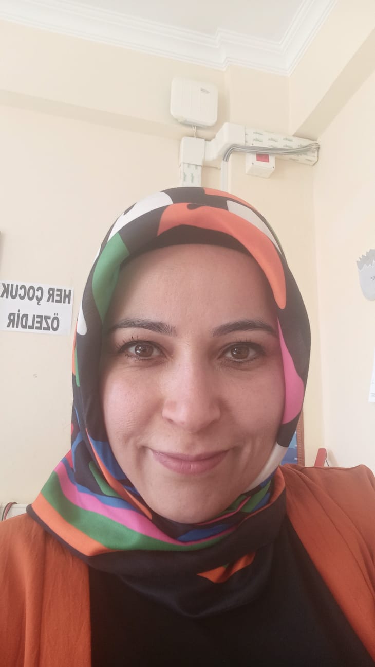 Renkli desenli bir başörtüsü ve turuncu bir ceket giymiş Asuman Düz’ün, sade açık renk bir arka plan önünde yakından çekilmiş görüntüsü.